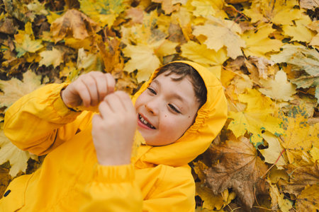Joyful Child in Yellow Raincoat Laying in Autumn Leaves at a Parkの写真素材