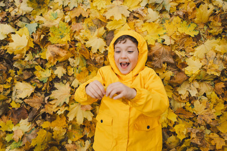 Joyful Child in Yellow Raincoat Laying in Autumn Leaves at a Park