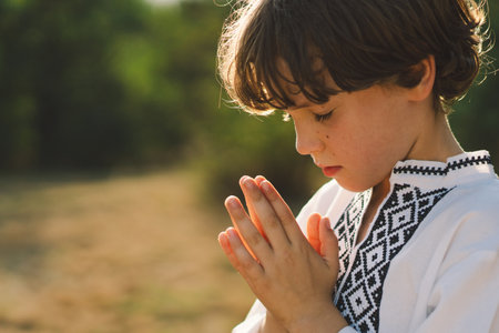A young child wearing a traditional embroidered shirt quietly prays outdoorsの写真素材