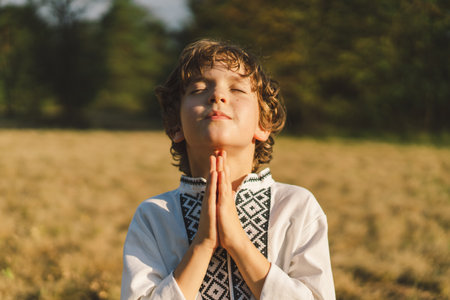 A young child wearing a traditional embroidered shirt quietly prays outdoorsの写真素材