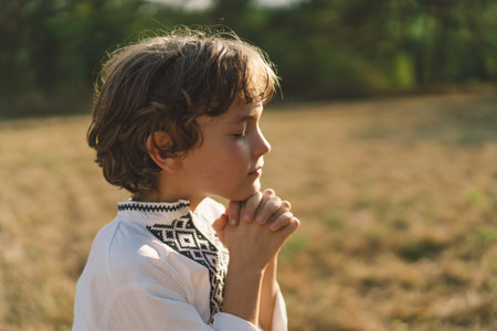 A young child wearing a traditional embroidered shirt quietly prays outdoorsの写真素材