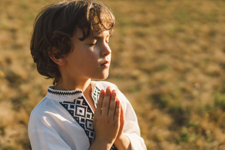 A young child wearing a traditional embroidered shirt quietly prays outdoorsの写真素材