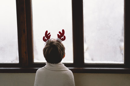 A child with reindeer antlers gazes out of a snowy window on a winter morningの写真素材