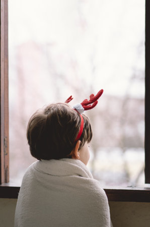 A child with reindeer antlers gazes out of a snowy window on a winter morningの写真素材