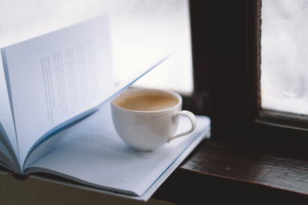 Cozy winter day with steaming coffee, open book, and candlelight by the snowy windowの写真素材