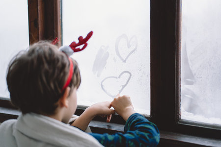 A child with reindeer antlers gazes out of a snowy window on a winter morningの写真素材