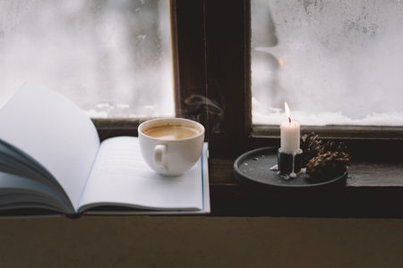 Cozy winter day with steaming coffee, open book, and candlelight by the snowy windowの写真素材