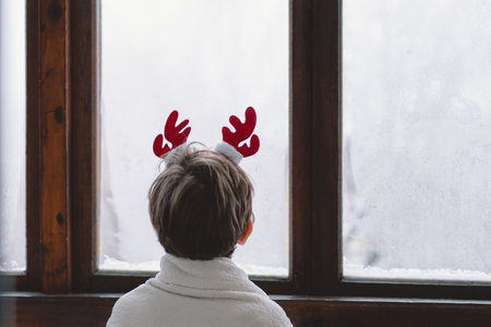 A child with reindeer antlers gazes out of a snowy window on a winter morningの写真素材
