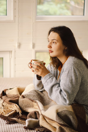 A woman is sitting in a cozy room, enjoying a coffee and reading a book during a relaxing morningの写真素材