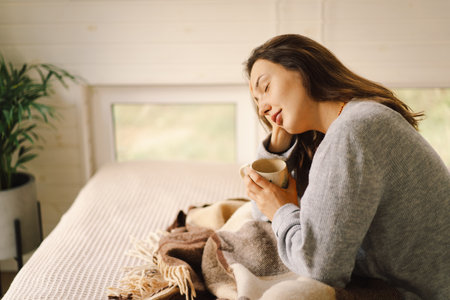 A woman is sitting in a cozy room, enjoying a coffee and reading a book during a relaxing morningの写真素材