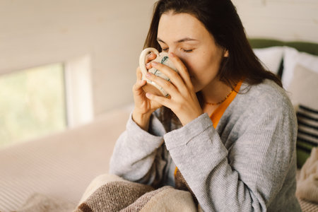 A woman is sitting in a cozy room, enjoying a coffee and reading a book during a relaxing morningの写真素材