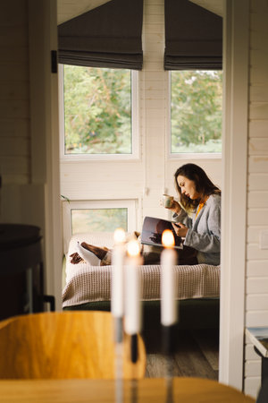 A woman is sitting in a cozy room, enjoying a coffee and reading a book during a relaxing morningの写真素材