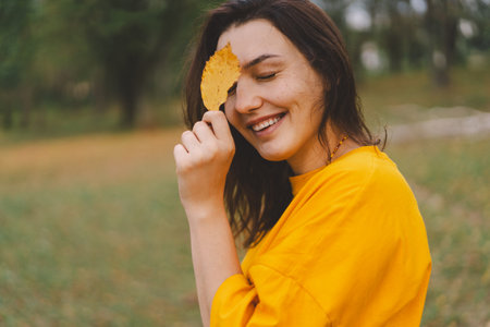 A woman enjoys a autumn day while playfully holding a yellow leaf