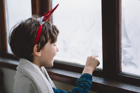A child with reindeer antlers gazes out of a snowy window on a winter morning