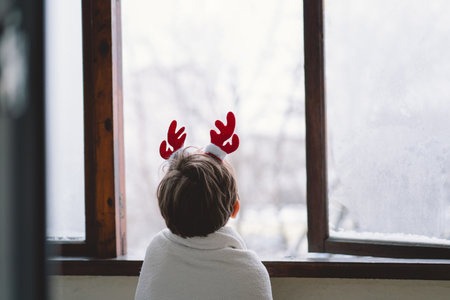 A child with reindeer antlers gazes out of a snowy window on a winter morningの写真素材