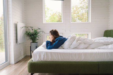 Joyful woman relaxing on soft white bed linen in a cozy bedroom during morning hoursの写真素材