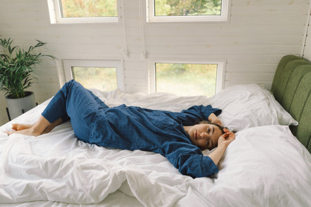 Joyful woman relaxing on soft white bed linen in a cozy bedroom during morning hoursの写真素材