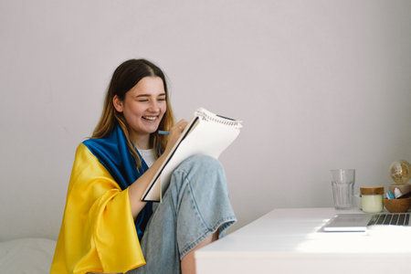 A Ukrainian teenage girl is wrapped in the Ukrainian flag and is drawing or doing homeworkの写真素材