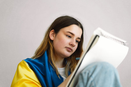 A Ukrainian teenage girl is wrapped in the Ukrainian flag and is drawing or doing homeworkの写真素材