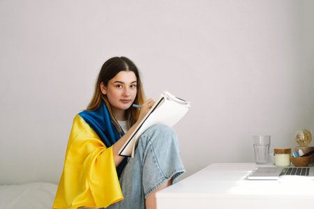 A Ukrainian teenage girl is wrapped in the Ukrainian flag and is drawing or doing homeworkの写真素材