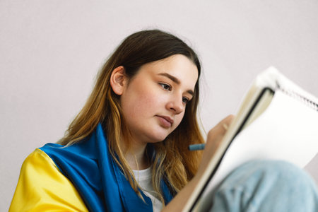 A Ukrainian teenage girl is wrapped in the Ukrainian flag and is drawing or doing homeworkの写真素材