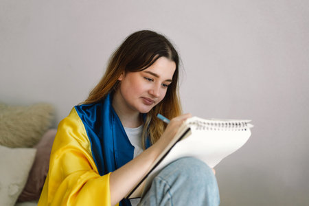 A Ukrainian teenage girl is wrapped in the Ukrainian flag and is drawing or doing homeworkの写真素材