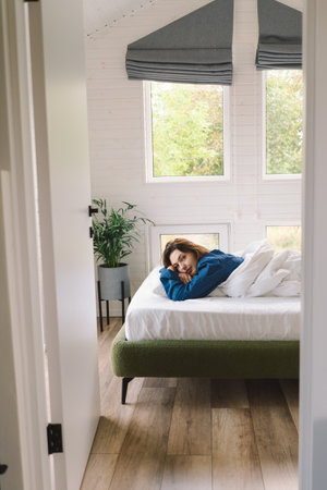 Joyful woman relaxing on soft white bed linen in a cozy bedroom during morning hoursの写真素材