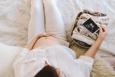 A pregnant woman lies on a bed with an ultrasound image and a basket of baby clothesの写真素材
