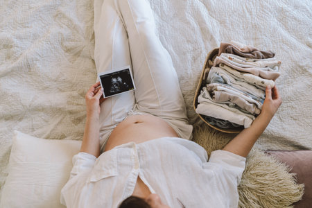 A pregnant woman lies on a bed with an ultrasound image and a basket of baby clothesの写真素材