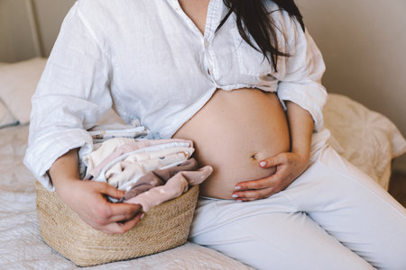 Expectant mother holds basket of baby clothes while gently touching her bellyの写真素材