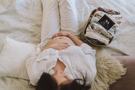 A pregnant woman lies on a bed with an ultrasound image and a basket of baby clothesの写真素材