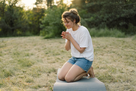 Woman closed her eyes, praying in a field during beautiful sunset. Hands folded in prayer concept for faithの写真素材