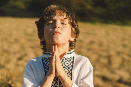 A young child wearing a traditional embroidered shirt quietly prays outdoors