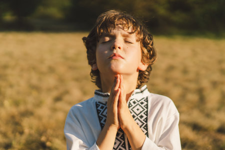 A young child wearing a traditional embroidered shirt quietly prays outdoors