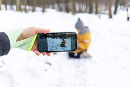 Child playing in snow captured on phone while adult watches in winter forest setting