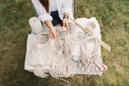 Woman crafts bag using macrame technique outdoors near tents. Outdoor hobbies.