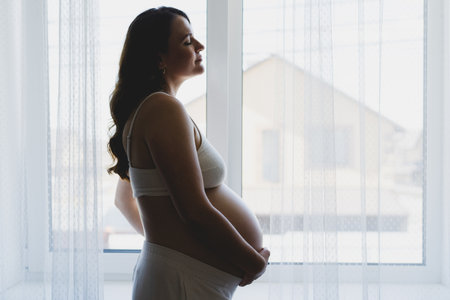 Pregnant woman smiling and touching her belly by a windowの写真素材