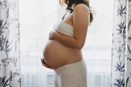 Pregnant woman smiling and touching her belly by a windowの写真素材
