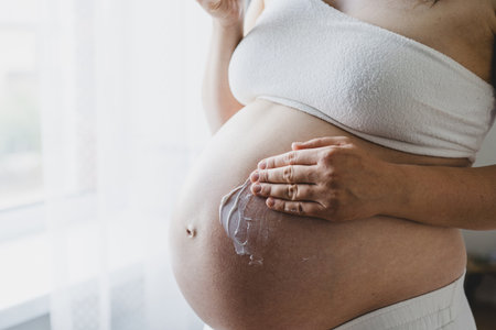 Pregnant woman applying cream on her belly while standing by a window in a bright roomの写真素材