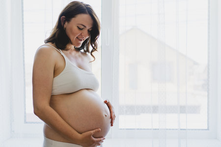 Pregnant woman smiling and touching her belly by a window