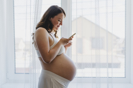 Expecting mother smiles while texting near window, enjoying quiet moment at home during pregnancyの写真素材