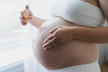 Pregnant woman applying cream on her belly while standing by a window in a bright roomの写真素材