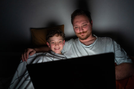Father and son watch a movie on a laptop while sitting on a bed at nightの写真素材