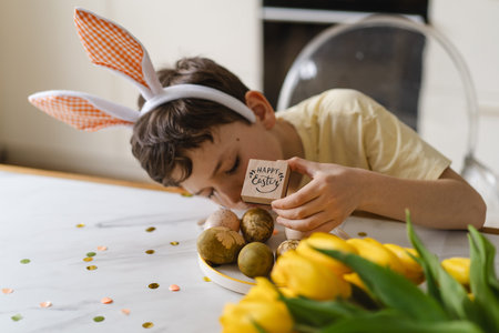 Smiling child wearing bunny ears with natural Easter eggs at home kitchenの写真素材