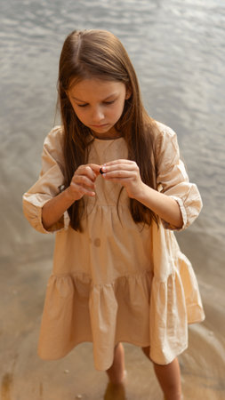 A girl in a beige dress with loose long hair stands barefoot in the water or river or sea, holds a shell or stone in her hands, examines it with interest, picking it with her fingerの写真素材