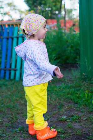 A little girl in a headscarf and rubber boots stands sideways, gestures with her hands, smiles and speaks. A child on the move on a farm or in a villageの写真素材