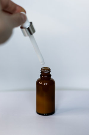 Brown dropper bottle without label, human hand holding pipette with white serum or cream. Cosmetic product on white background, front view.の写真素材
