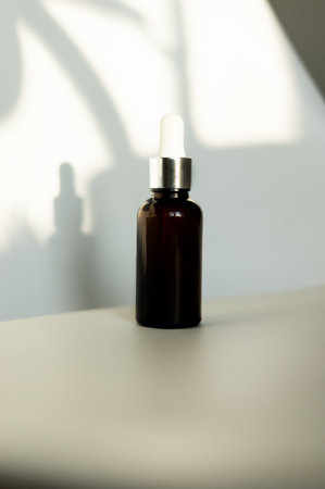 Brown dropper bottle or glass jar without label. Cosmetic product mockup on white background illuminated by bright sunlight from window, front view.の写真素材