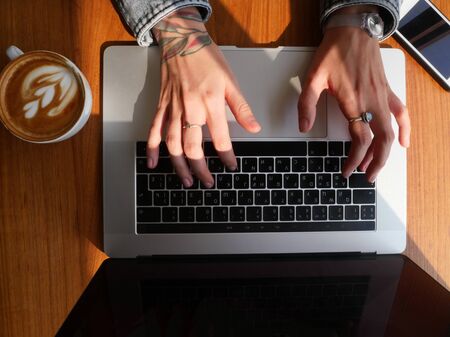 Top view tattooed girl hands texting on laptop drinking coffee.