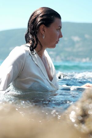 Woman in focus in black bikini and white wet shirt behind unfocused cliff in turquoise water...